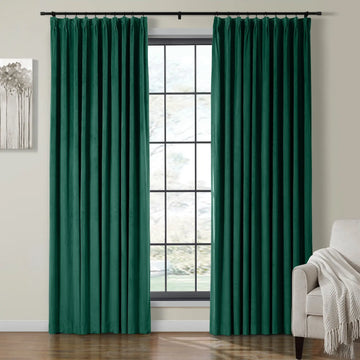 Two luxurious dark emerald green velvet curtains on a black rod frame a grid-pane window with a subtle outdoor blur, complemented by an abstract grey tree painting and a beige armchair with a cream fringed throw