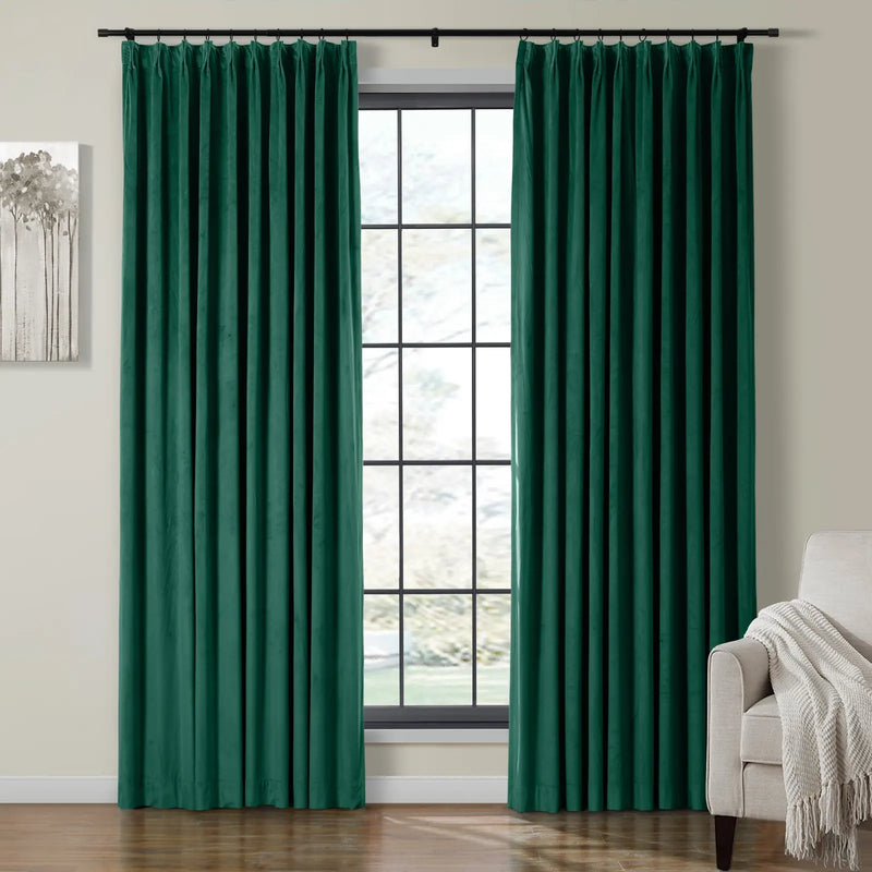 Two luxurious dark emerald green velvet curtains on a black rod frame a grid-pane window with a subtle outdoor blur, complemented by an abstract grey tree painting and a beige armchair with a cream fringed throw