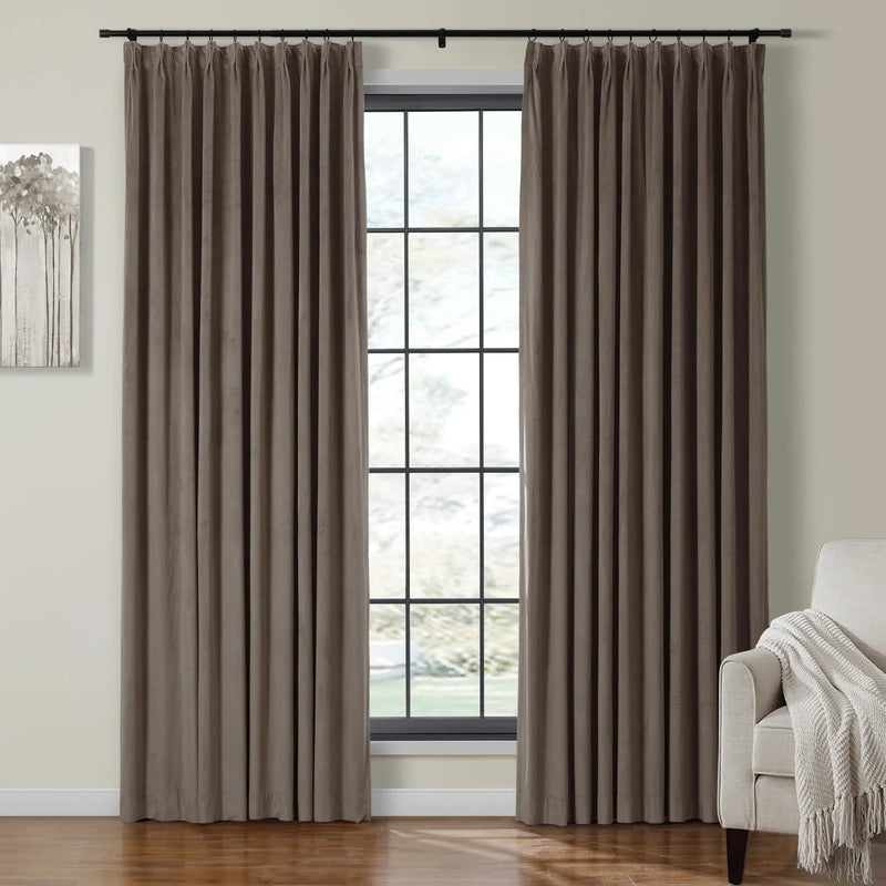 Plush taupe brown velvet pleated curtains hanging from a black rod frame a multi-pane window, beside an abstract gray tree artwork and a cream armchair with a fringed throw blanket