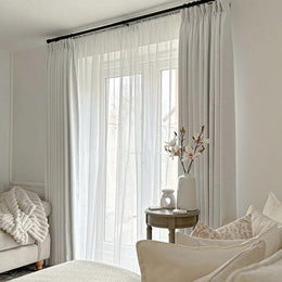 Interior photo of a bright bedroom showcasing a large window dressed with white sheer and light grey blackout curtains on a black rod, complemented by a small round table holding a white vase with delicate white and pink flowers and a ceramic oil burner
