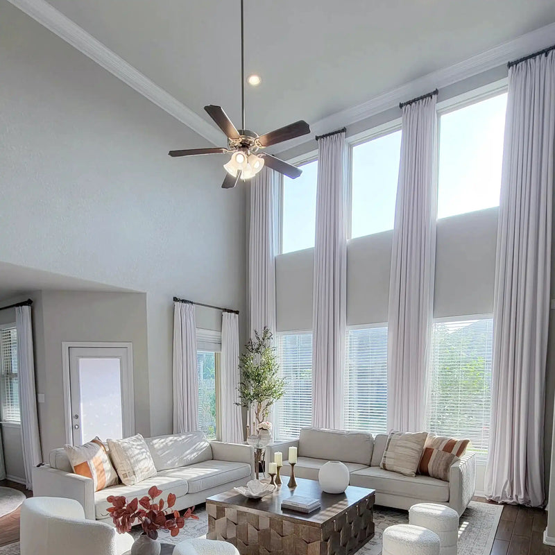 A spacious modern living room showcasing a high two-story ceiling with a dark ceiling fan, tall white curtains framing large sunlit windows, and two white sofas adorned with patterned neutral and orange-toned throw pillows centered around a textured geometric wooden coffee table