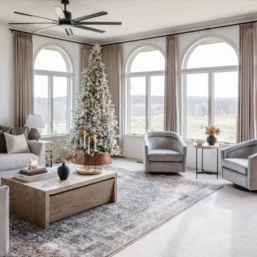 A festive modern living room with a large flocked Christmas tree adorned with gold and white ornaments, featuring three expansive arched windows overlooking a serene winter landscape, accented by gray velvet swivel chairs and a black ceiling fan