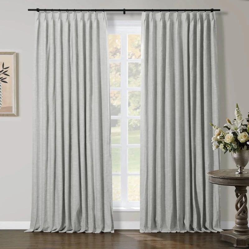 Light gray pleated curtains on a black rod in a room with dark wood floors and a visible window, complemented by a carved side table with a bouquet of white and yellow flowers and framed abstract plant art