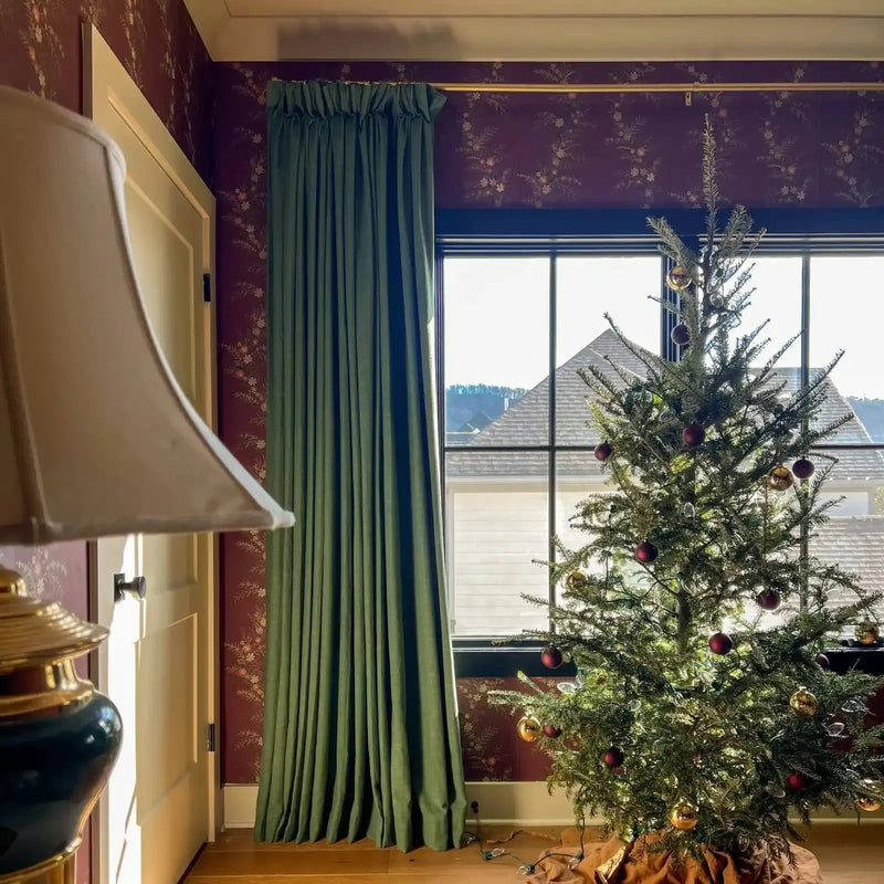 An indoor Christmas scene featuring a sparse tree adorned with red and gold ornaments, set against a window revealing residential rooftops, framed by deep green curtains and rich burgundy floral wallpaper