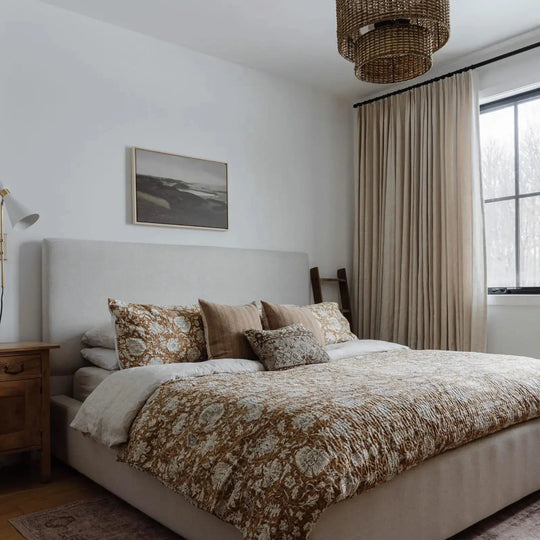 Photograph of a cozy bedroom featuring a large bed with an earthy floral patterned quilt, a distinctive woven tiered chandelier, and elegant light beige linen curtains