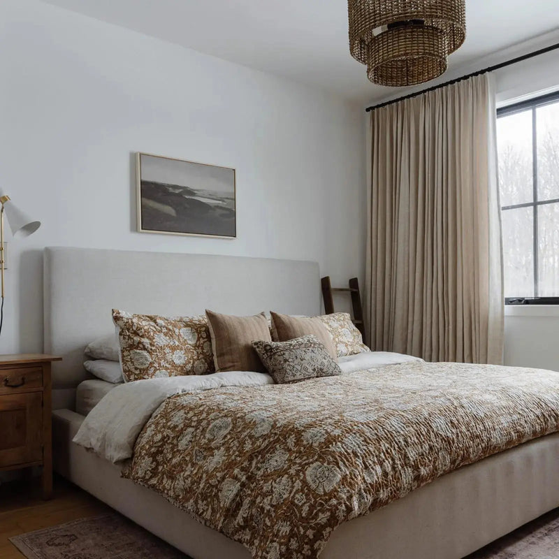 Photograph of a cozy bedroom featuring a large bed with an earthy floral patterned quilt, a distinctive woven tiered chandelier, and elegant light beige linen curtains