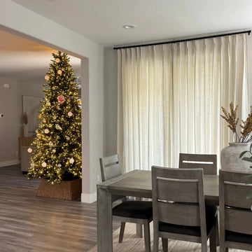 Interior view of a modern dining room featuring a gray wooden table and chairs with dark cushions, looking into a living room with a brightly lit Christmas tree adorned with white, gold, and a unique red handprint ornament, framed by tall off-white curtains and a large gray ceramic vase with dried branches