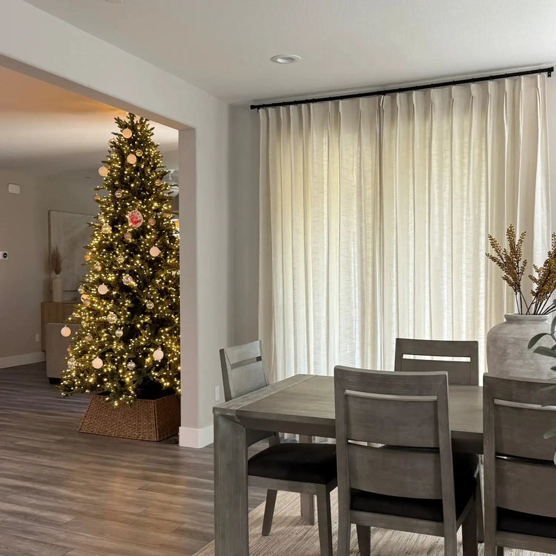 Interior view of a modern dining room featuring a gray wooden table and chairs with dark cushions, looking into a living room with a brightly lit Christmas tree adorned with white, gold, and a unique red handprint ornament, framed by tall off-white curtains and a large gray ceramic vase with dried branches