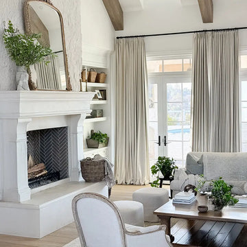 A bright, neutral-toned living room featuring a grand white stone fireplace with dark herringbone tile, an ornate gold mirror, exposed wooden ceiling beams, and French doors opening to a view of a backyard pool, adorned with natural textures and abundant greenery