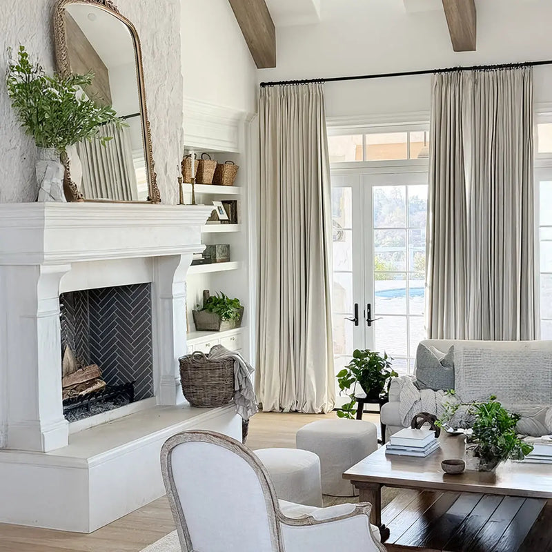 A bright, neutral-toned living room featuring a grand white stone fireplace with dark herringbone tile, an ornate gold mirror, exposed wooden ceiling beams, and French doors opening to a view of a backyard pool, adorned with natural textures and abundant greenery