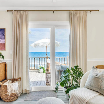 Vibrant coastal interior looking out through glass doors onto a wooden deck with a white fringed umbrella and bright blue ocean, showcasing an indoor Monstera plant and woven basket