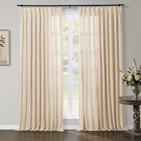 Natural linen beige pinch-pleat curtains frame a multi-pane window, complemented by a dark wood twisted-base table with a silver vase holding white and green flowers