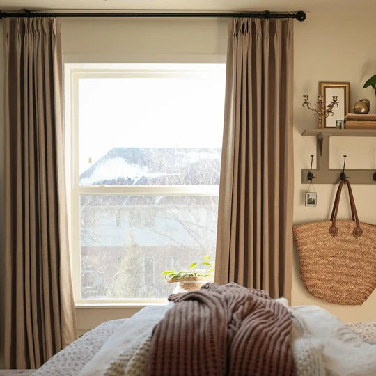 A cozy bedroom interior with beige drapes framing a window overlooking a snowy roof, featuring a woven straw tote bag hanging on the wall and a knitted rust-colored throw blanket on the bed