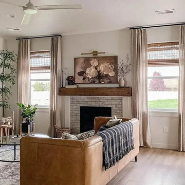 Interior photograph of a cozy modern living room with a tan leather sofa draped in a plaid throw, a light brick fireplace featuring a rustic wood mantel and abstract floral wall art, flanked by large windows with natural linen curtains and bamboo shades