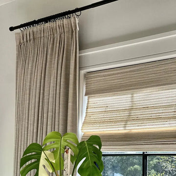 A bright interior window scene featuring beige textured drapes on a black rod, a woven Roman blind, and a vibrant variegated Monstera plant with large split green leaves