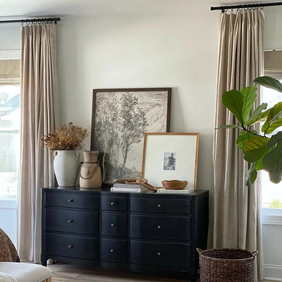 A black dresser styled with a large framed tree sketch, a white vase of dried flowers, and a vibrant fiddle leaf fig plant in a bright room with neutral linen curtains