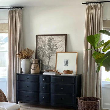 A black dresser styled with a large framed tree sketch, a white vase of dried flowers, and a vibrant fiddle leaf fig plant in a bright room with neutral linen curtains