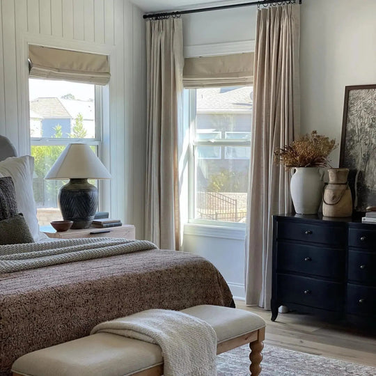 A neutral bedroom interior with white shiplap walls a richly patterned brown bedspread a textured dark bronze lamp on a light wood nightstand and a black dresser displaying a white vase of dried botanicals