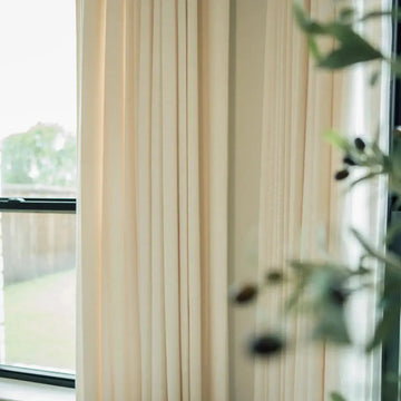 Photograph of an interior window framed by cream-colored pleated curtains, offering a glimpse of a green lawn and wooden fence outside, with a blurred green leafy plant adorned with dark berries partially obscuring the view