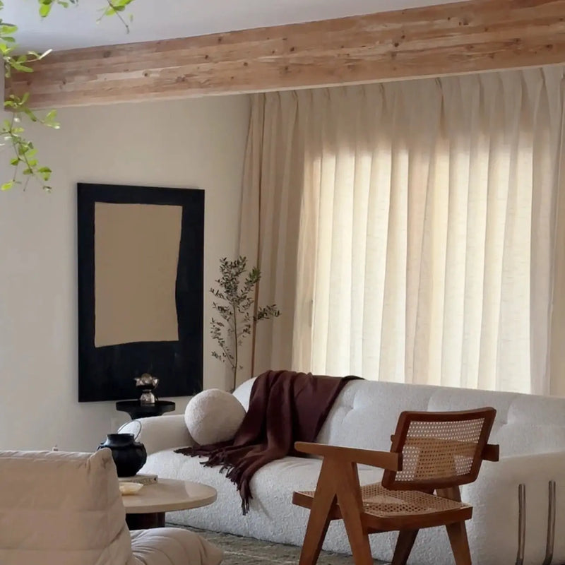 A stylish minimalist living room featuring a white bouclé sofa with a dark brown throw, a large abstract black and beige artwork, and a prominent cane and wood chair under a wooden beam ceiling