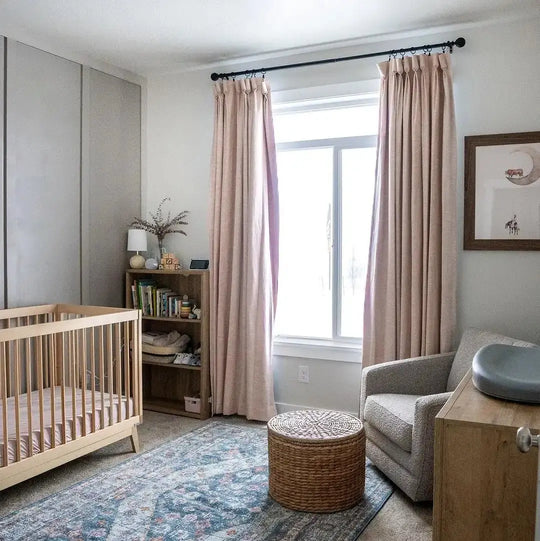 Photograph of a modern baby nursery featuring a light wood crib blush pink linen curtains a blue and pink patterned rug and a woven round ottoman