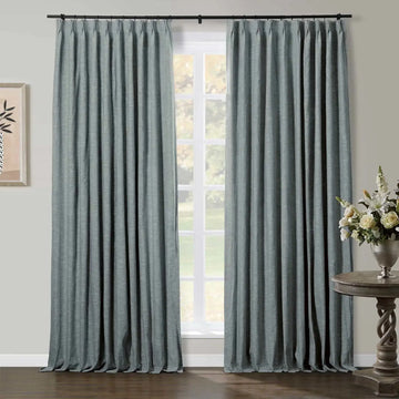 Teal blue pinch pleat linen curtains framing a white paned window in a domestic setting with a dark wood floor a distressed wood side table holding a silver vase with white and cream flowers and a framed botanical art print
