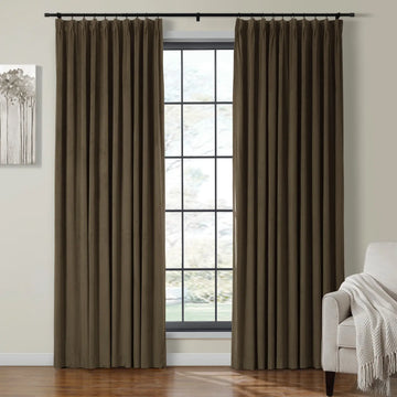 Dark brown pleated blackout curtains on a black rod frame a multi-pane window with a blurred green outdoor view in a modern interior featuring a cream armchair with a white woven throw and a gray abstract tree painting