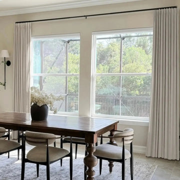 Photograph of an elegant dining room interior featuring a dark wooden dining table with ornate turned legs, modern chairs with light upholstered seats, white pleated curtains framing large windows overlooking lush green trees and a black metal railing, and a white floral centerpiece in a dark vase