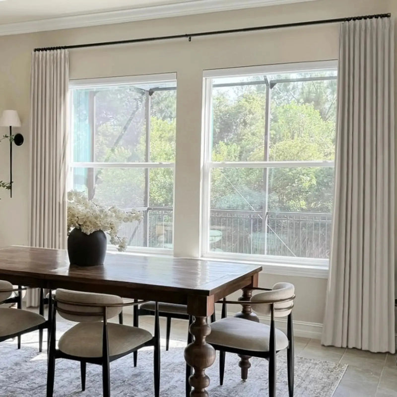 Photograph of an elegant dining room interior featuring a dark wooden dining table with ornate turned legs, modern chairs with light upholstered seats, white pleated curtains framing large windows overlooking lush green trees and a black metal railing, and a white floral centerpiece in a dark vase