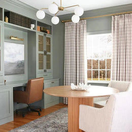 An inviting home office showcasing sage green built-in cabinetry, a rich brown leather desk chair, a natural wood fluted table with a white coral-like bowl, and a sleek brass globe chandelier