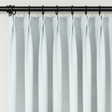 Photograph of light blue-grey textured pinch pleat curtains on a black rod with rings
