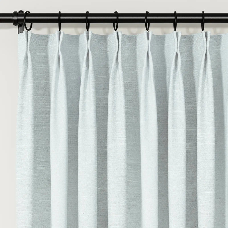 Photograph of light blue-grey textured pinch pleat curtains on a black rod with rings