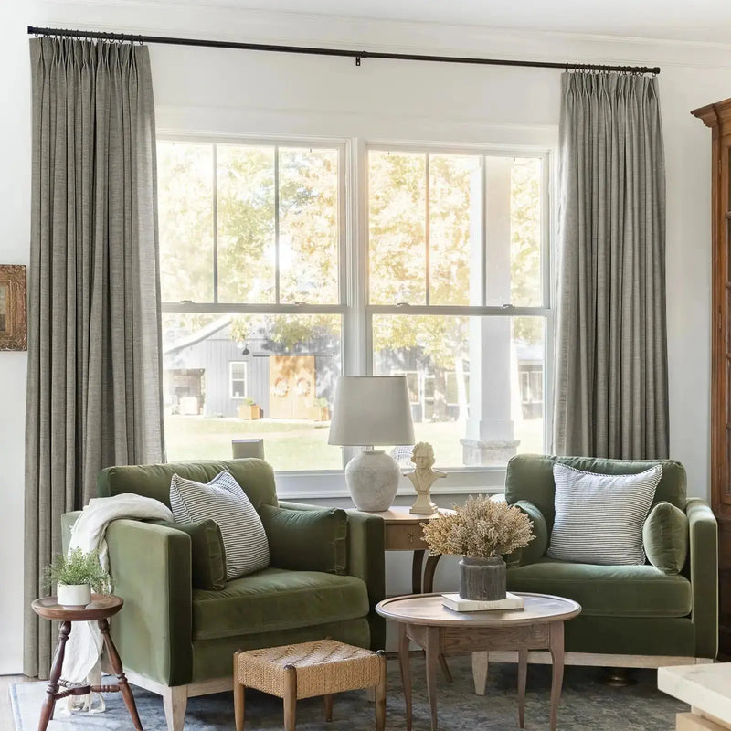 Photograph of a cozy living room interior featuring two plush olive green velvet armchairs with striped pillows, a classical bust on a side table, and a bright view through large windows of a grey barn outside