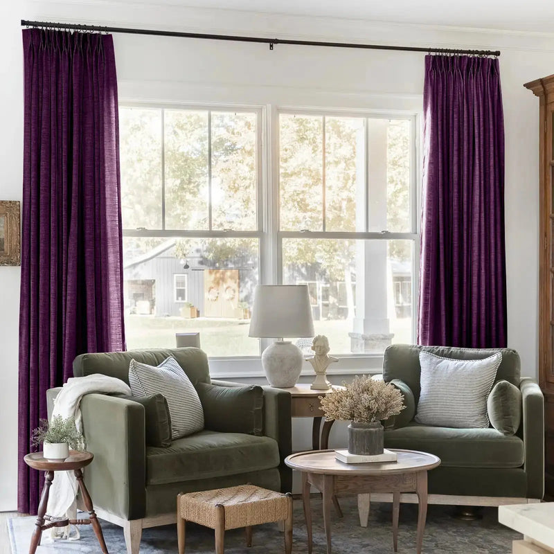 A warm and inviting interior shot featuring two olive green armchairs with striped pillows, flanked by rich plum velvet curtains, a white bust sculpture, and a woven ottoman, looking out onto a bright green yard with a grey barn and white porch column