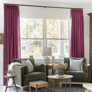 A cozy living room interior features plum colored velvet curtains framing a large window with a distant gray barn, two olive green velvet armchairs with striped throw pillows, and a classical bust sculpture on a small table