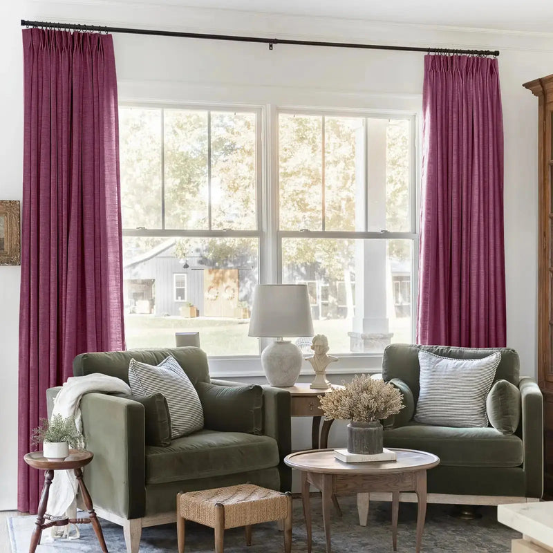 A cozy living room interior features plum colored velvet curtains framing a large window with a distant gray barn, two olive green velvet armchairs with striped throw pillows, and a classical bust sculpture on a small table