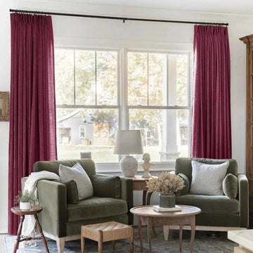 Cozy living room with olive-green velvet armchairs and striped cushions, magenta pinch-pleat curtains framing a large window overlooking a rustic barn, featuring a white classical bust and textured ceramic lamp