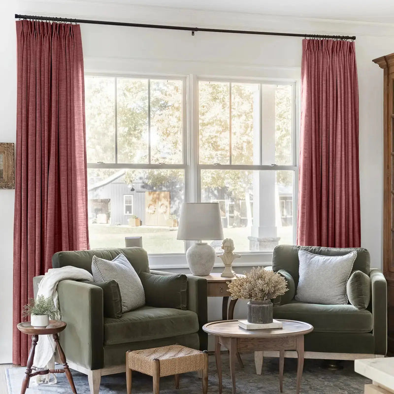 Interior of a charming living room with vibrant cranberry pleated draperies flanking a window revealing a rustic gray barn, complemented by two plush olive green velvet armchairs and striped throw pillows