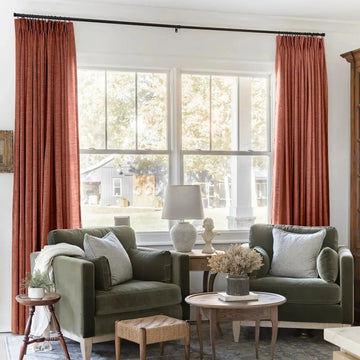 A cozy living room interior with burnt orange pinch-pleat curtains framing a window overlooking a rustic barn and white porch columns, featuring two sage green velvet armchairs with striped pillows and a wicker ottoman