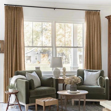 Cozy living room interior with two olive green velvet armchairs, tall tan pleated curtains, and a view through large windows to a white porch column and rustic barn, featuring striped throw pillows and a classical bust statue