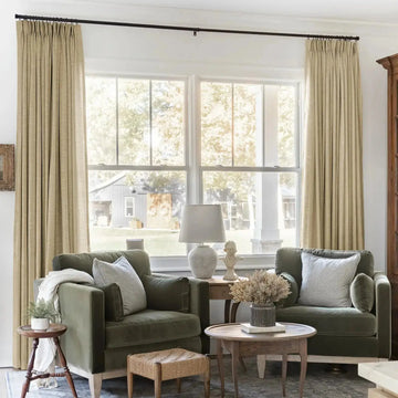 A cozy living room interior with two olive green velvet armchairs, striped pillows, a white classical bust statue, and floor-length beige linen curtains, offering a view of a serene rural backyard with a rustic barn featuring two wreaths