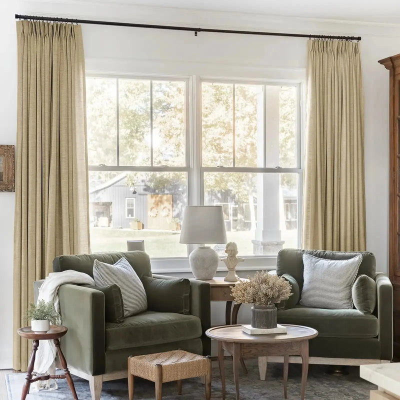 A cozy living room interior with two olive green velvet armchairs, striped pillows, a white classical bust statue, and floor-length beige linen curtains, offering a view of a serene rural backyard with a rustic barn featuring two wreaths