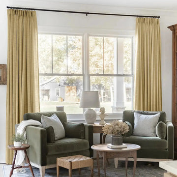 A bright, inviting living room with two olive green velvet armchairs, a woven jute ottoman, and gold pleat curtains framing a window overlooking a rustic barn with decorative wreaths