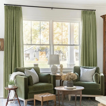 Interior design photograph of a cozy living room featuring two olive green velvet armchairs with striped throw pillows, light green curtains, and a classical bust sculpture, all framed by large white windows with visible exterior columns overlooking a rustic barn adorned with two decorative wreaths