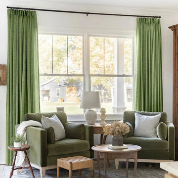 Photograph of a cozy living room interior featuring two olive green velvet armchairs, bright green pinch-pleat curtains, a classic bust sculpture, and a woven footstool, all set against a large window overlooking a rustic country barn and white porch pillar
