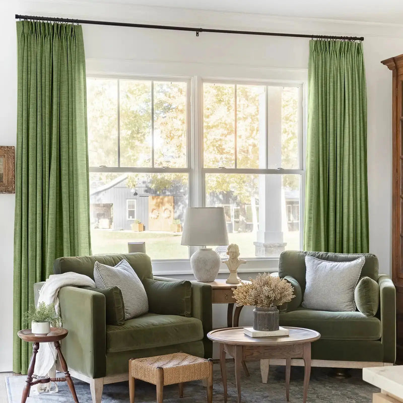 Photograph of a cozy living room interior featuring two olive green velvet armchairs, bright green pinch-pleat curtains, a classic bust sculpture, and a woven footstool, all set against a large window overlooking a rustic country barn and white porch pillar