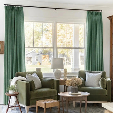 A cozy living room interior featuring two olive green velvet armchairs, tall green pleated drapes, and a sunny window view revealing a grey barn adorned with two wreaths