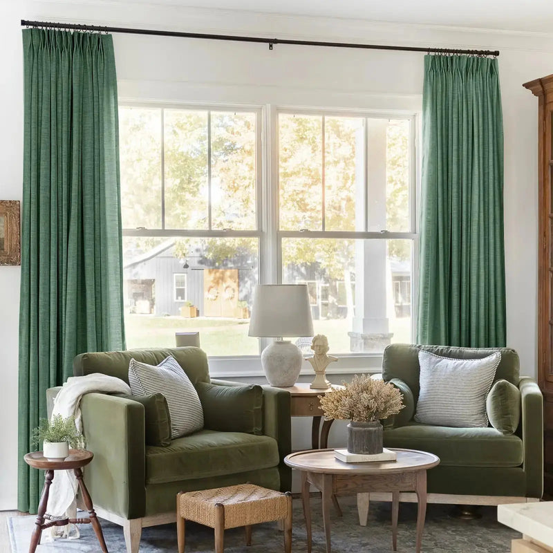 A cozy living room interior featuring two olive green velvet armchairs, tall green pleated drapes, and a sunny window view revealing a grey barn adorned with two wreaths