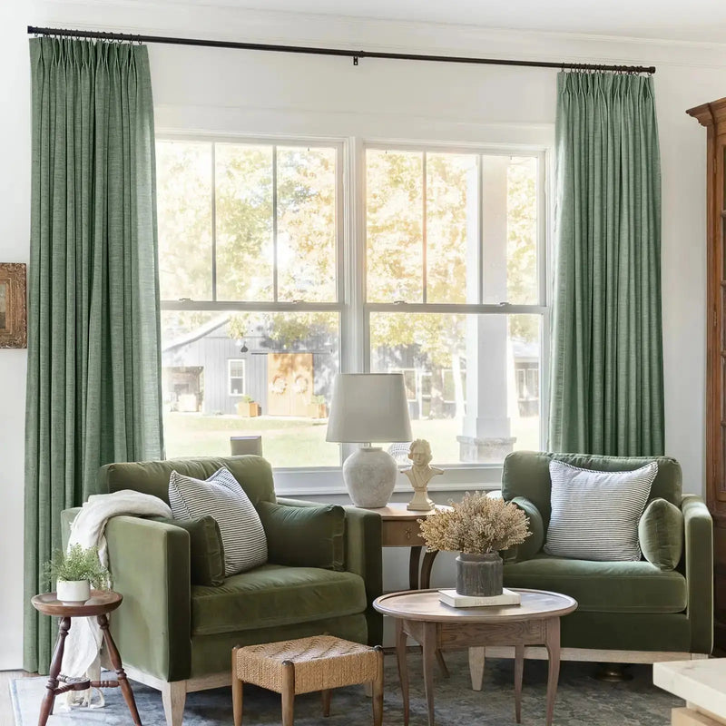 A stylish interior living space with two olive green velvet armchairs, striped throw pillows, a woven rattan stool, and a bust sculpture on a side table, offering a bright window view of a rustic barn and sun-dappled trees