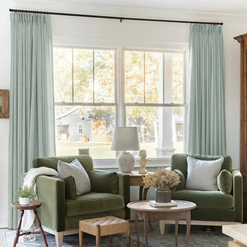 A warm interior view of a living room with two olive green velvet armchairs, light mint green pinch-pleat curtains framing a large window, a white classical bust on a side table, and a rustic barn visible outside